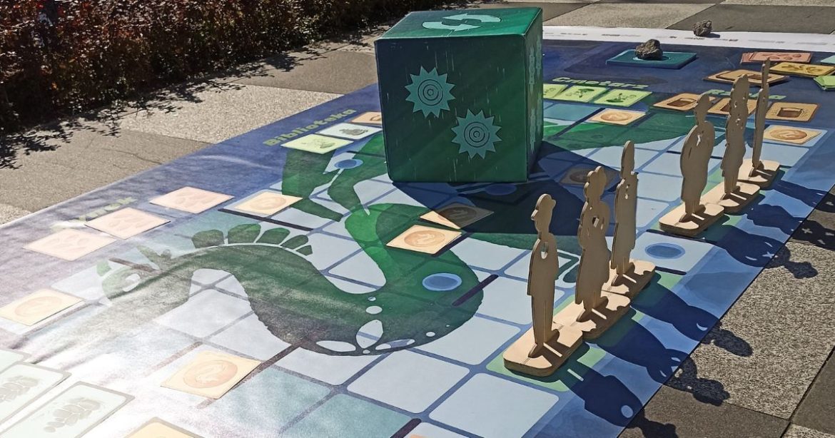 Planszowa gra wielkoformatowa rozłożóna na chodniku - na planszy stoją drewniane pionki i ogromna kostka.