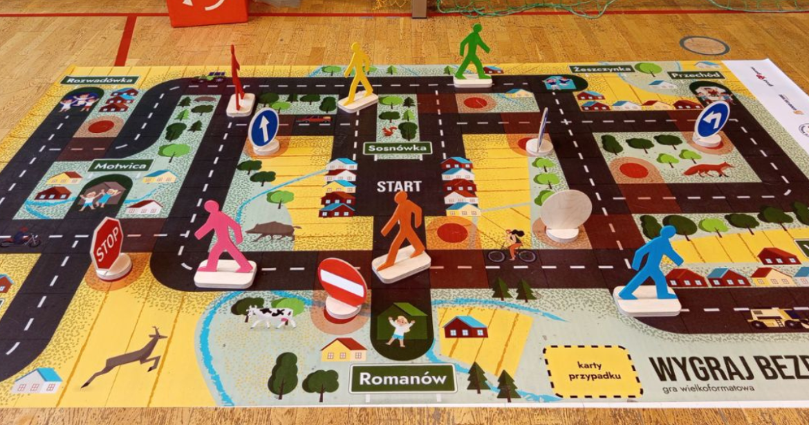 Na obrazku widać planszę wielkoformatowej gry planszowej skierowanej do dzieci. Jej celem jest promowanie Bezpieczeństwa Ruchu Drogowego.