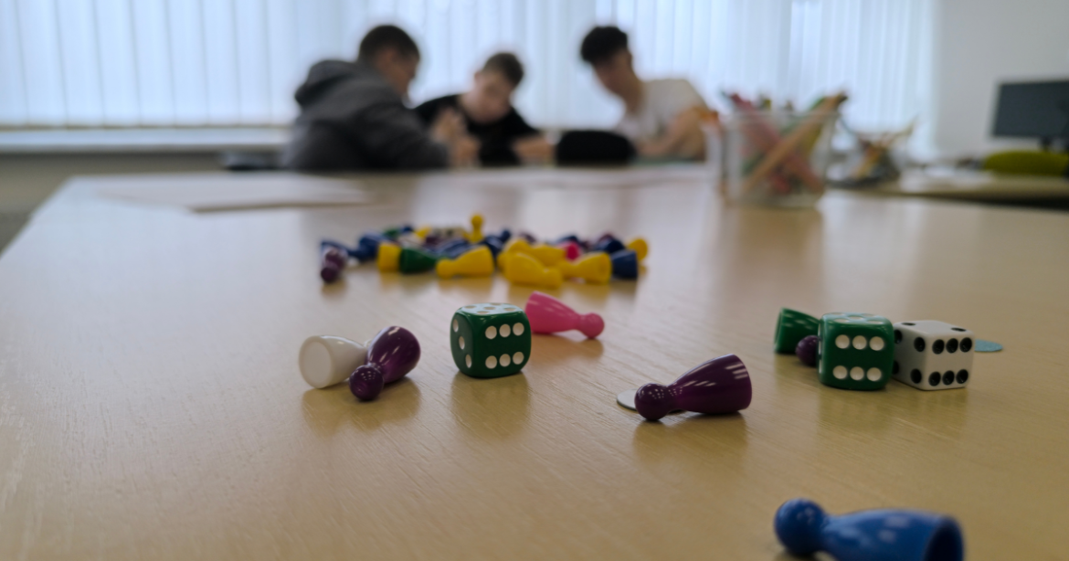Warsztaty tworzenia gier w Słupcy. Na pierwszym planie rozrzucone pionki i kostki do gry. W tle widać młodzież projektującą własną grę wielkoformatową.