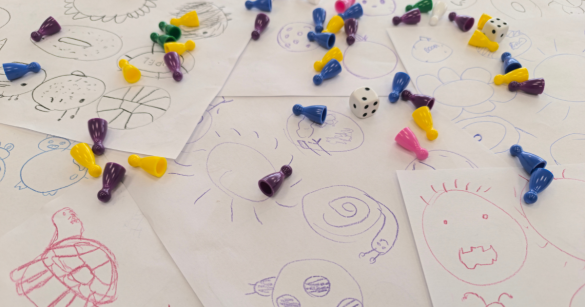 Pionki i kostki do gry rozrzucone po rysunkach stworzonych na warsztatach w Słupcy
